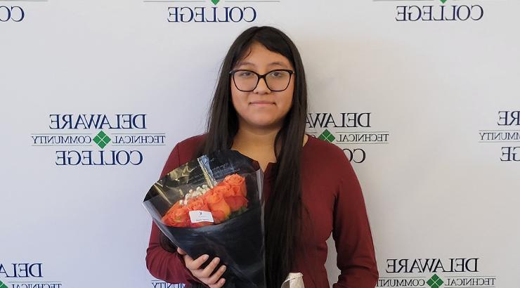 Breanna Vicente Huinac holding a bouquet of roses while standing in front of a white backdrop featuring the Delaware Tech logo in a tiled pattern